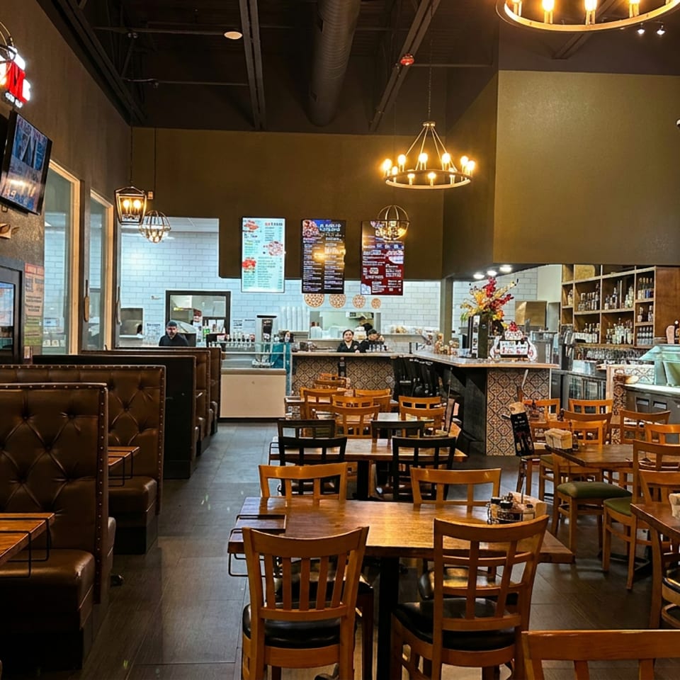 Spacious casual restaurant interior with leather booths, wooden tables, bar seating, chandeliers, and visible kitchen counters.