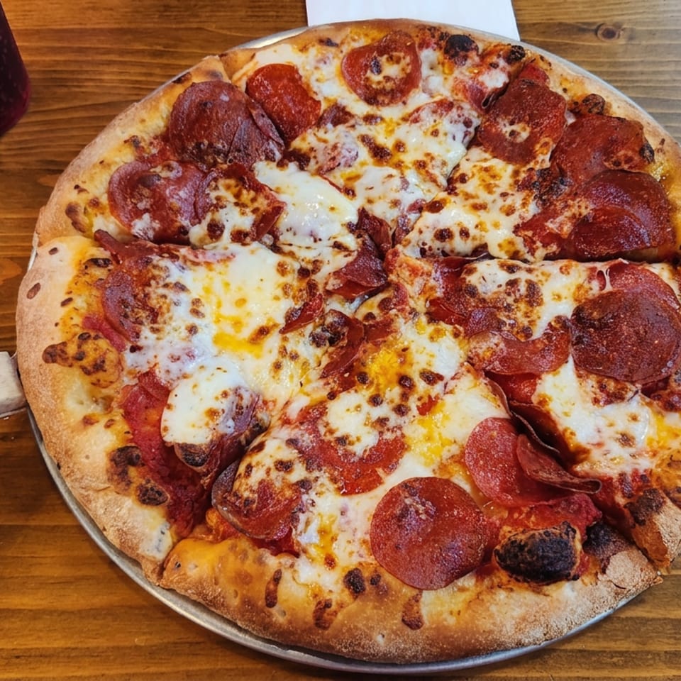 Pepperoni pizza with melted, browned cheese on a metal tray atop a wooden table, accompanied by a dark beverage.