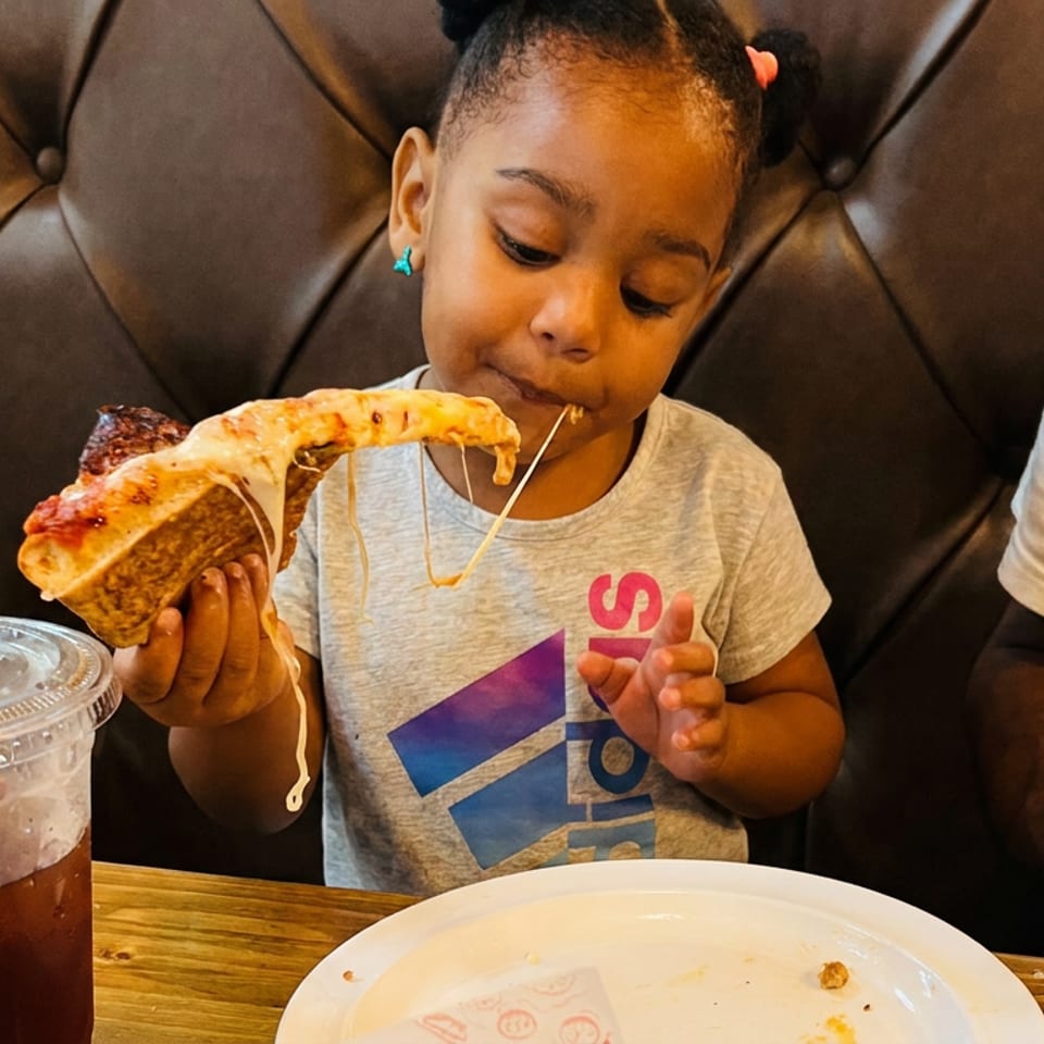 Child enjoys a cheese pizza slice with stretching mozzarella at a restaurant table beside an iced drink and adult.